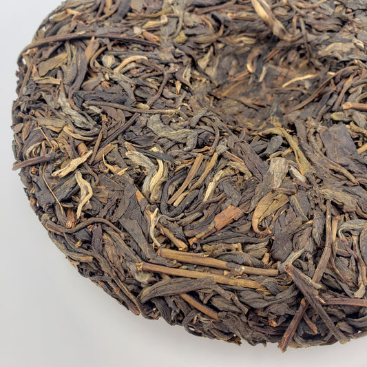 A close-up of a round compressed tea cake, showing the textured blend of dark brown, greenish, and light tan tea leaves, photographed on a plain white background.