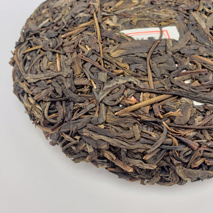 A close-up of a round compressed cake of dark brown tea leaves, showing the textured surface, with a small white label partially visible near the top edge, photographed on a plain white background.