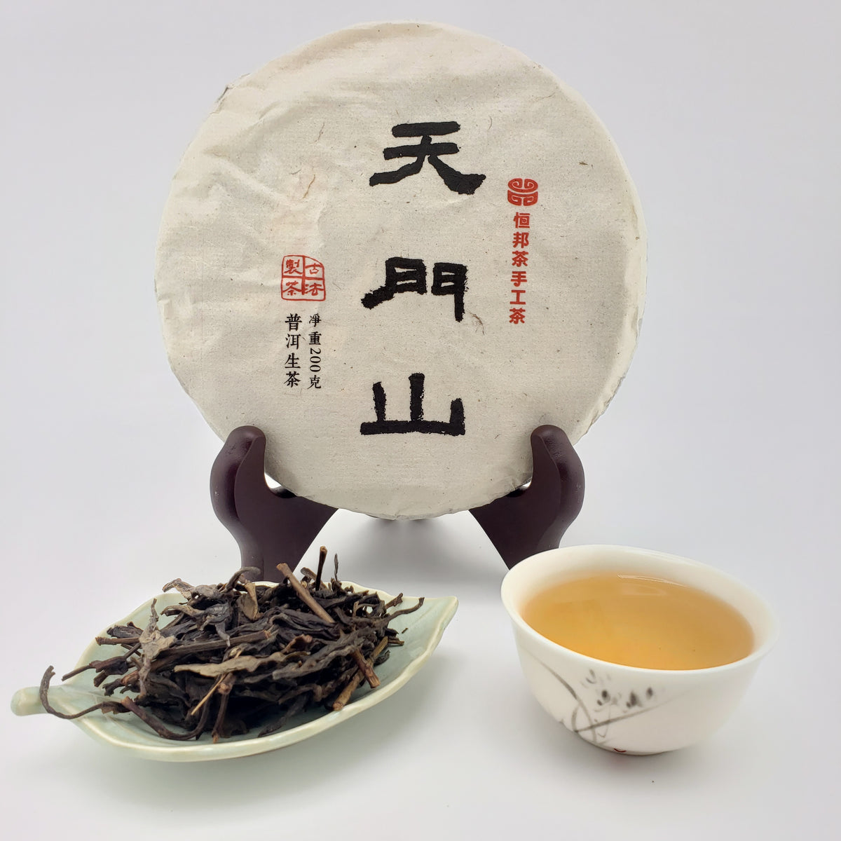 A round wrapped tea cake displayed upright on a stand, with a small dish of loose tea leaves and a cup of brewed tea on a white background. The tea cake has black and red Chinese calligraphy characters on the wrapping.