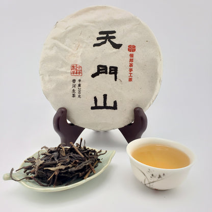 A round wrapped tea cake displayed upright on a stand, with a small dish of loose tea leaves and a cup of brewed tea on a white background. The tea cake has black and red Chinese calligraphy characters on the wrapping.