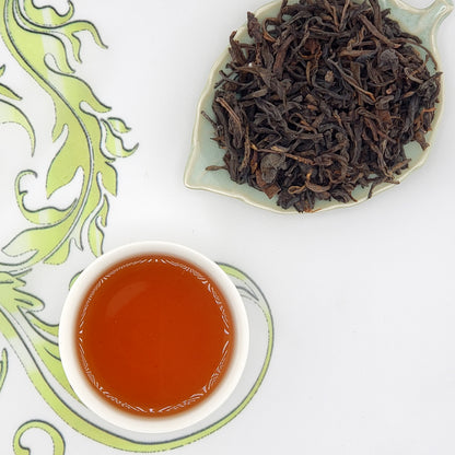 Dried tea leaves on a leaf-shaped plate with a cup of brewed tea on a decorative white and green background.