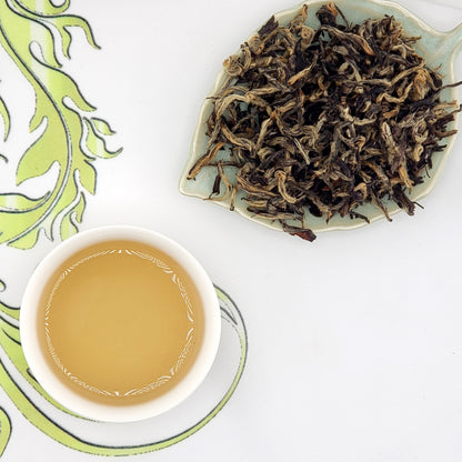 Dried tea leaves on a small plate next to a cup of brewed tea on a decorative mat.