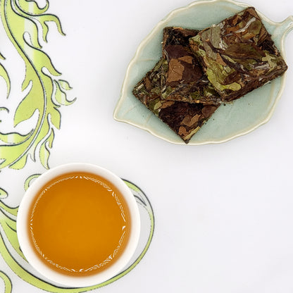 Tea leaves on a leaf-shaped plate with a cup of tea on a decorative mat.