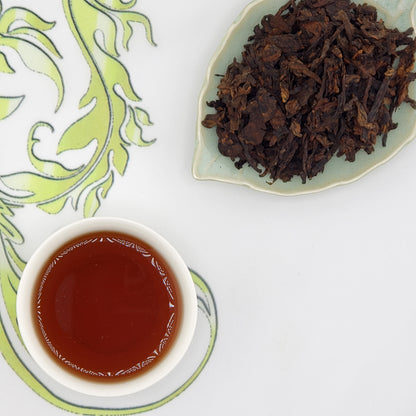 Dried tea leaves on a plate with a cup of brewed tea on a decorative mat.