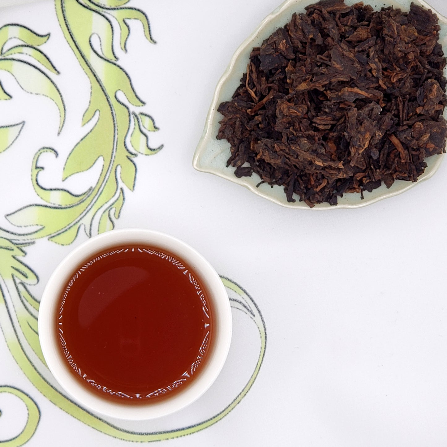 Cup of black tea and a dish of tea leaves on a decorative white and green surface.