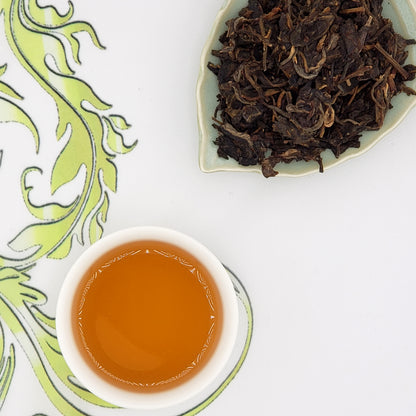 Dried tea leaves on a white plate with a cup of brewed tea on a decorative white surface.