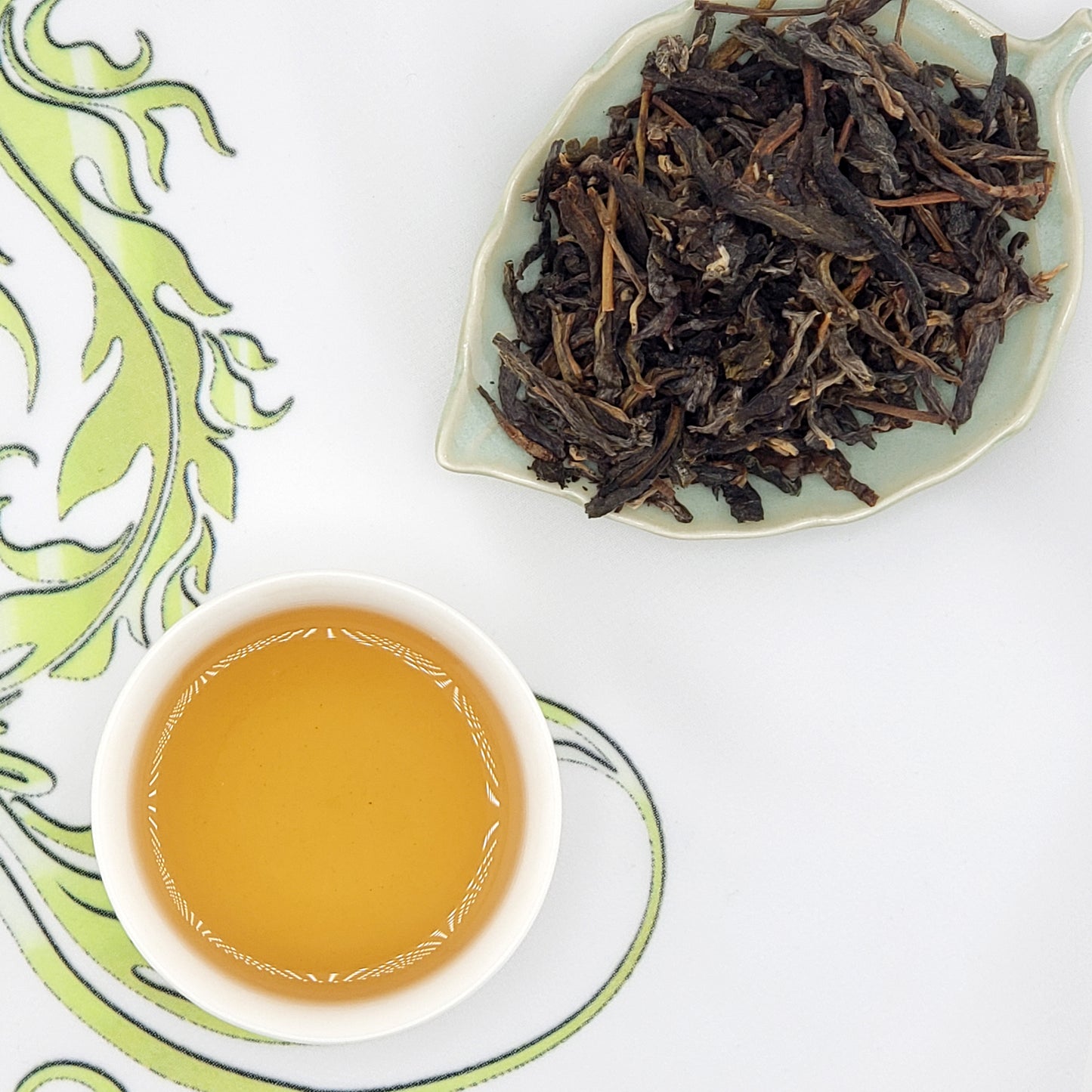 Dry tea leaves on a leaf-shaped dish with a cup of brewed tea on a decorative white background.