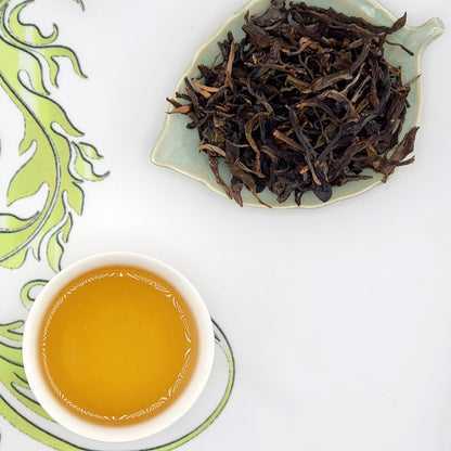 Dried tea leaves on a leaf-shaped plate with a cup of brewed tea on a decorative mat.