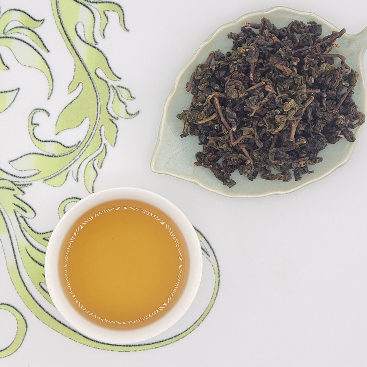 A cup of brewed honey-colored tea next to a dish of tea leaves, green and brown, and tightly rolled.