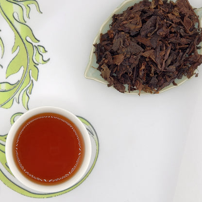 Dried tea leaves on a plate with a cup of brewed tea on a decorative mat.