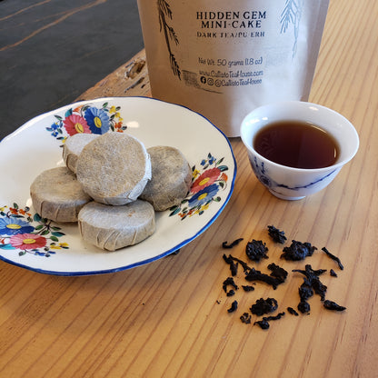 Tea cup with tea leaves, wrapped tea coins on a decorative plate, and tea packaging on a wooden surface.