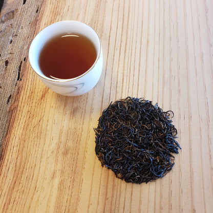 A small white cup of amber-brown tea sits on a light wood table beside a small pile of dark, curly loose-leaf tea.