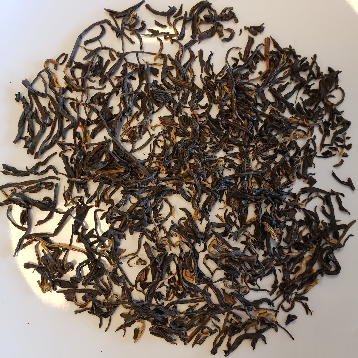 Dried loose-leaf tea leaves, primarily dark brown with lighter golden-brown tips, scattered on a plain white surface.