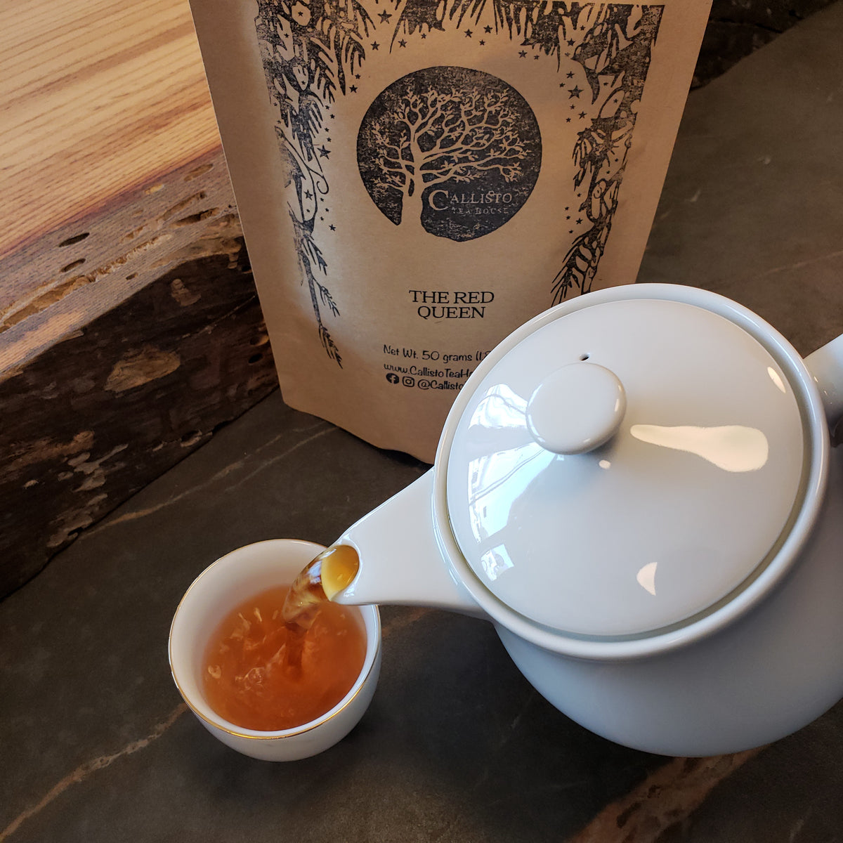A white ceramic teapot with a lid is shown pouring amber-colored tea into a small white cup, with a branded paper pouch labeled 'The Red Queen' standing behind it on a dark stone surface.