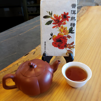 Tribute pu-erh tea brick with floral design packaging, yixing clay pot, white cup with tea, on a wooden surface.