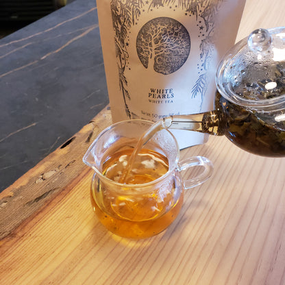 Glass teapot pouring tea into a clear glass mug with a package of White Pearls tea nearby.