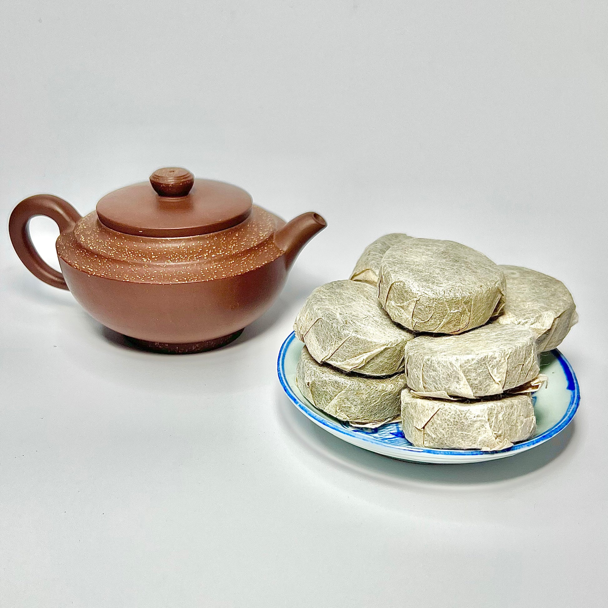 Ceramic teapot next to a plate of wrapped tea on a white background