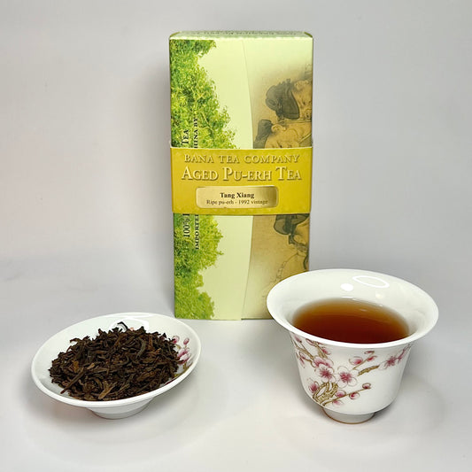 Tea packaging, cup with tea, and bowl of tea leaves on a gray background